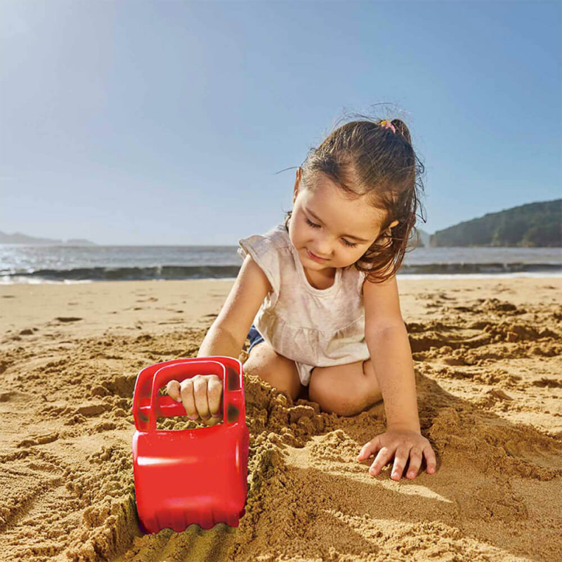 Petite fille à la plage avec sa pelleteuse manuelle rouge hape